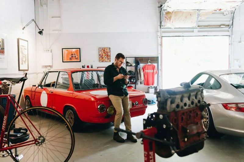 Man looking at phone while leaning up against a classic red BMW, with a silver Porsche and exposed engine also visible in a modern garage.