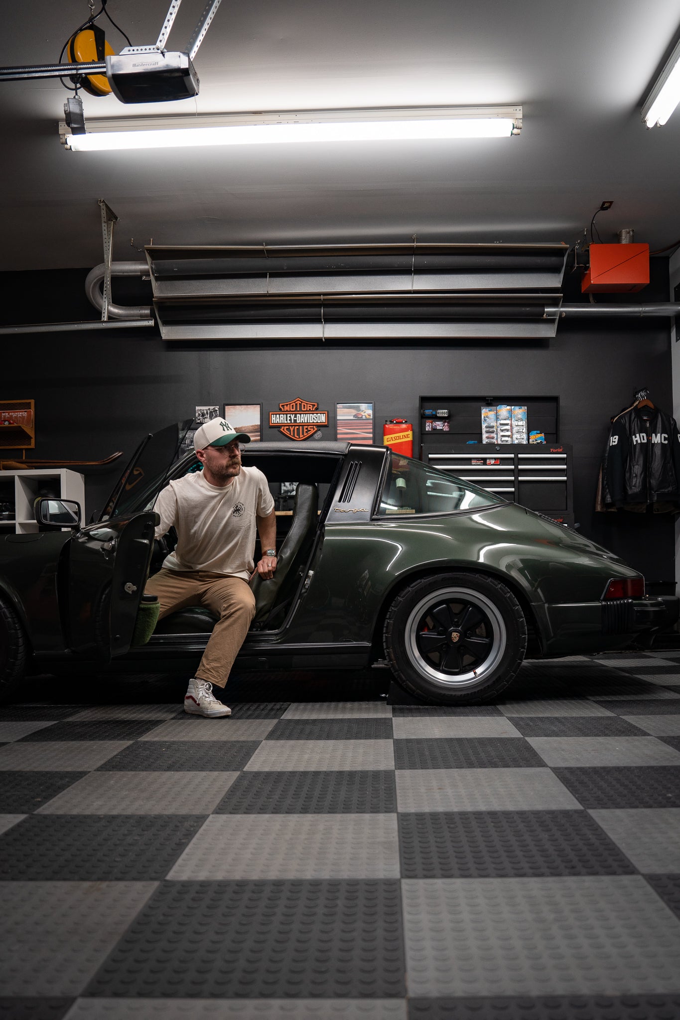 Owner sitting in a dark green 1977 Porsche 911S Targa inside a black-walled enthusiast garage