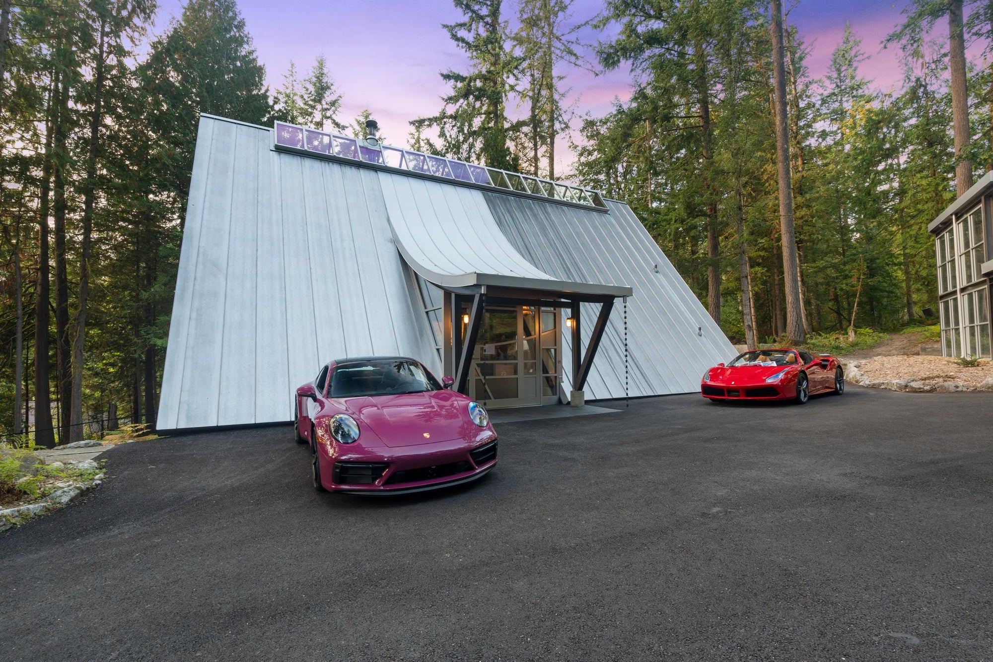 Modern A-Frame architecture with a Porsche and Ferrari parked in front, surrounded by trees.