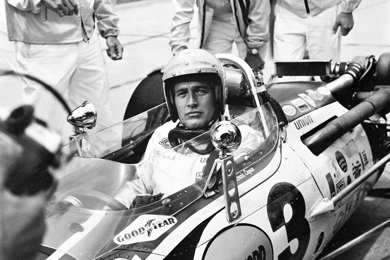 Paul Newman in a racing car with 'Goodyear' branding, surrounded by other people and cars, photo in black and white. 