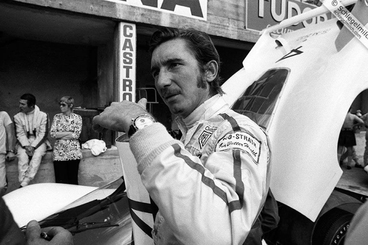 Black and white photo of a Jo Stiffert, a Formula one race car driver in a garage with his car and a Heuer watch on his wrist. 