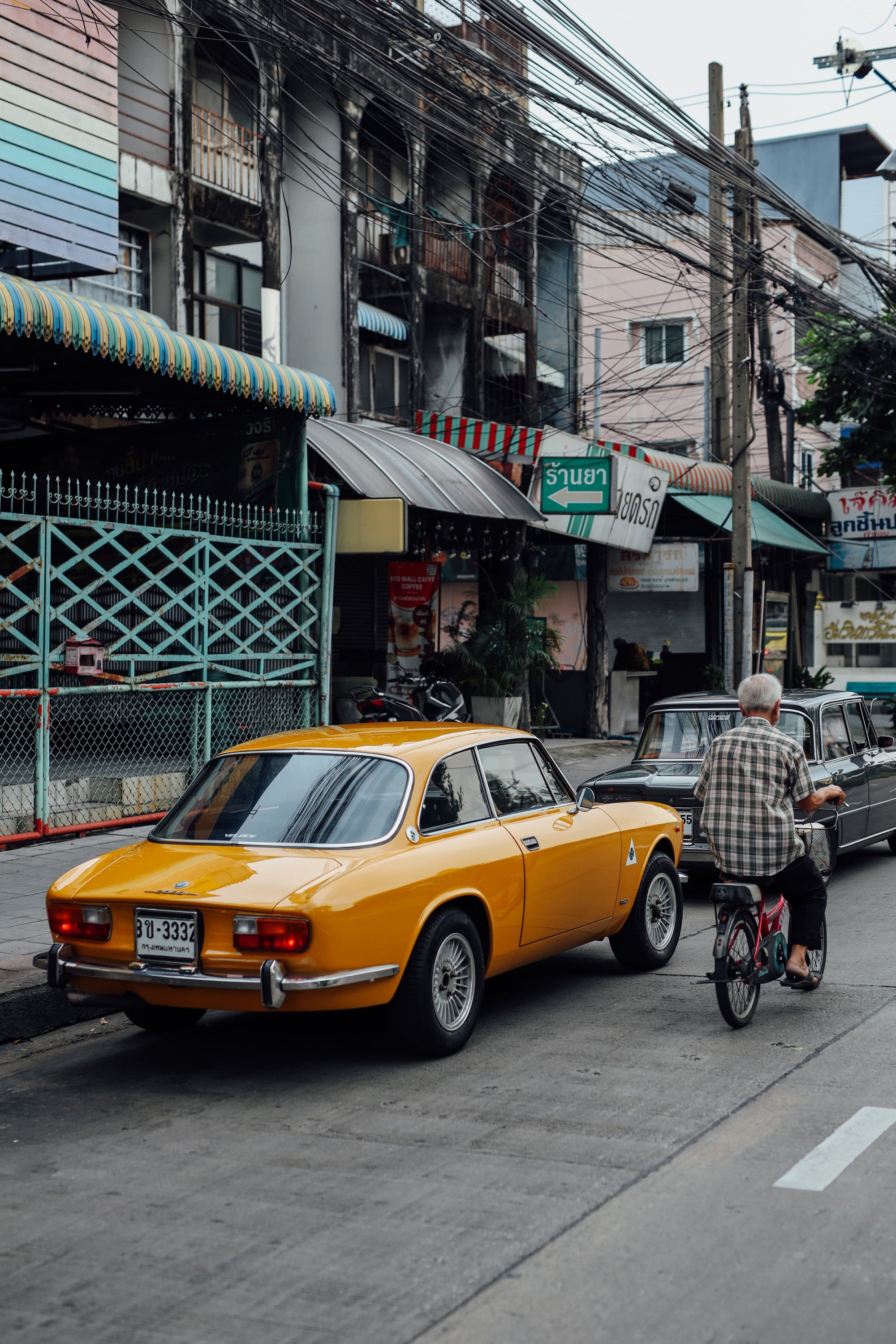 Rear view of a bright yellow vintage Alfa Romeo coupe parked on a Bangkok street, with an old man on a bicycle and tangled power lines overhead.