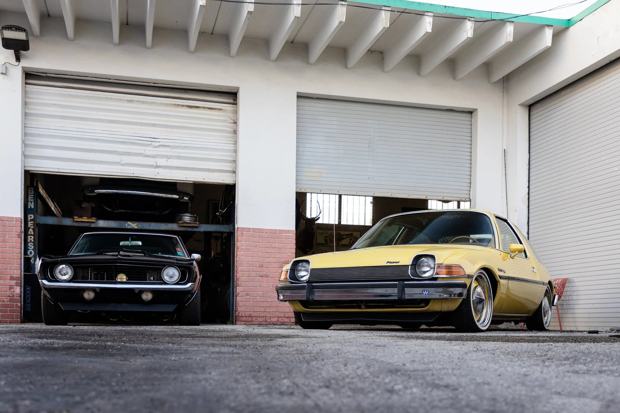 1969 Chevrolet Camaro and vintage yellow AMC Pacer parked in front of collector garage with a white building in the background.