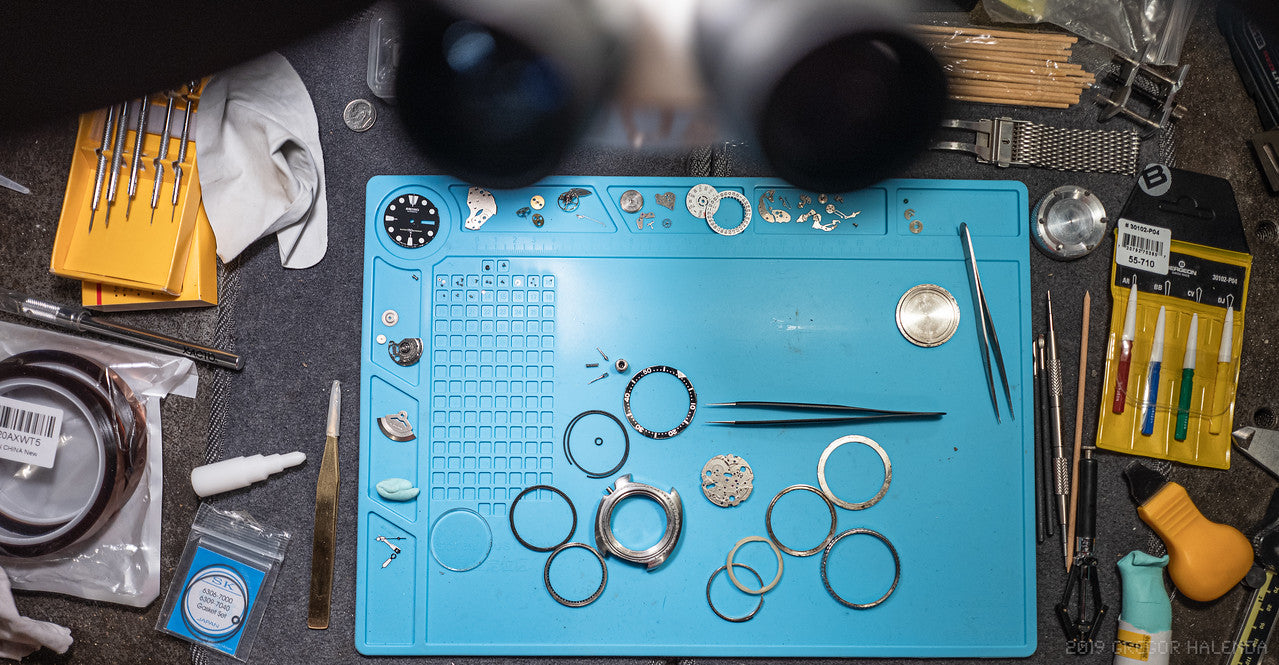 Blue work mat with watch components and tools on a dark surface on top of a workbench. 