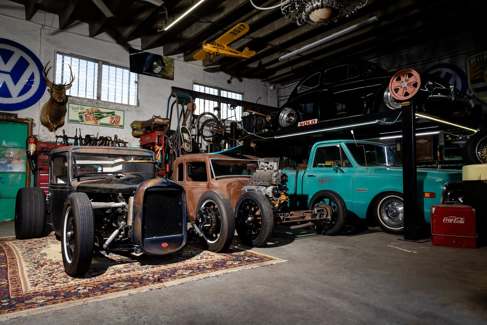 Two custom hot rods, a GMC truck, and a black VW beetle parked on a patterned rug in a Ft. Lauderdale garage with vintage decor.