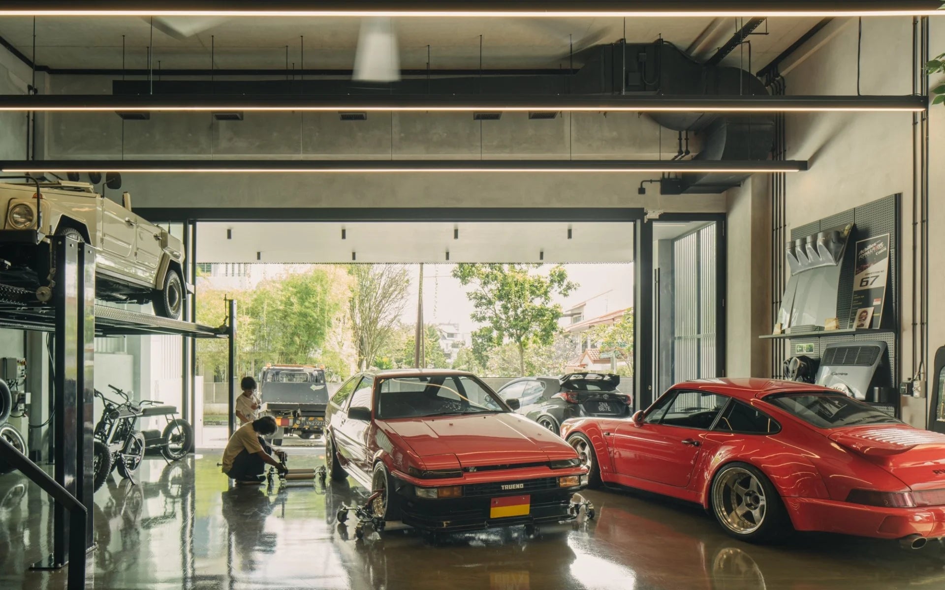 Modern garage interior with red Porsche 911, Toyota AE86, VW Thing, and mechanics working.