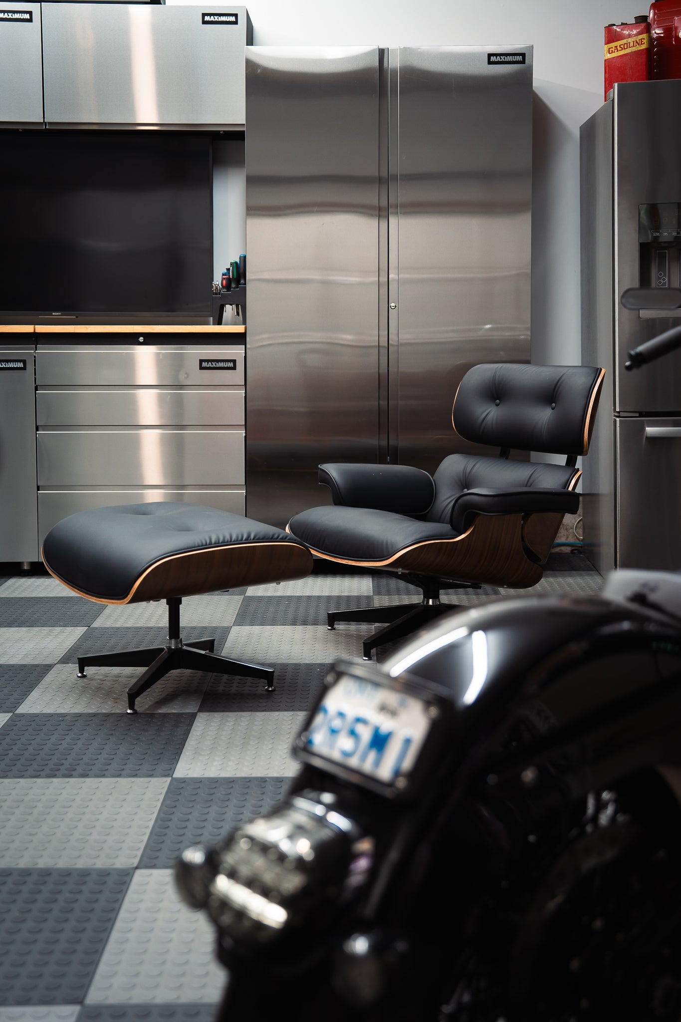 Modern garage interior with stainless steel workbench and cabinets and a black leather Eames chair with ottoman, motorcycle in foreground.