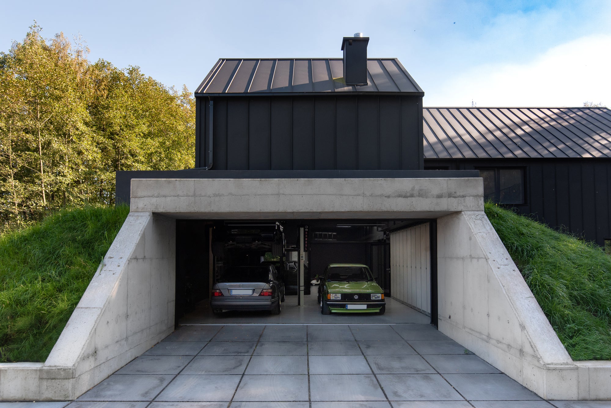 Garage integrated into hillside with exposed concrete entry beneath modern home with classic Mercedes, BMW, and VW golf inside 