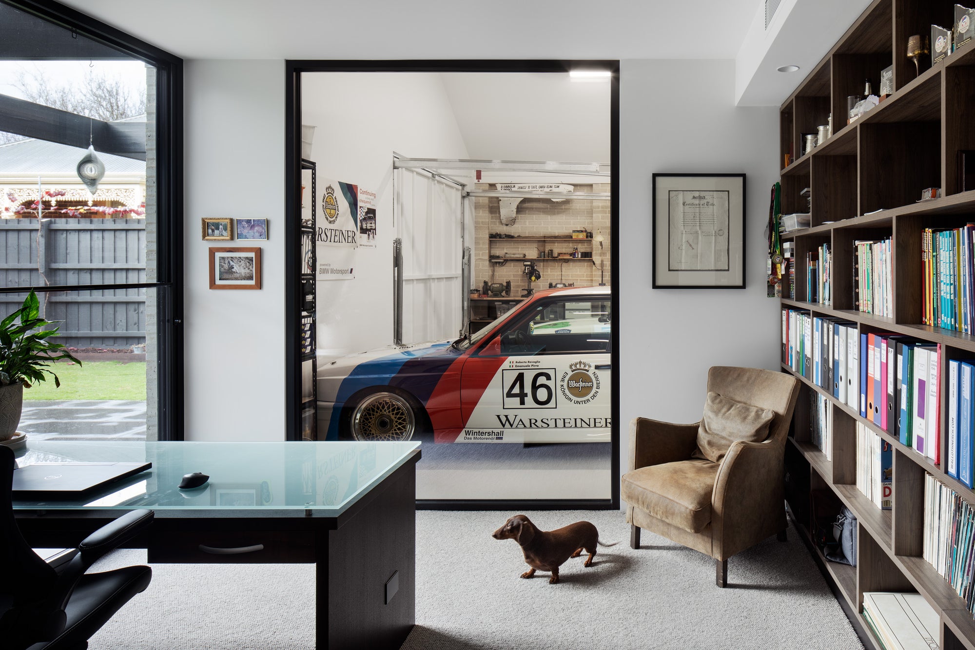 Home office picture-frame window framing a BMW Warsteiner race car in BMW Motorsport livery in the adjacent workshop