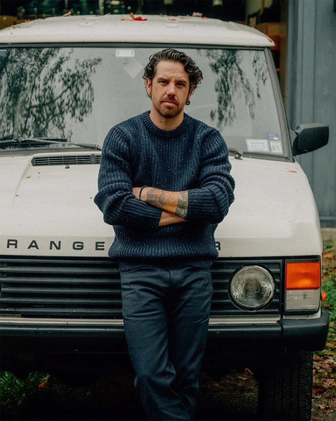 Man in navy sweater and dark jeans stands in front of a vintage white Range Rover with arms crossed.