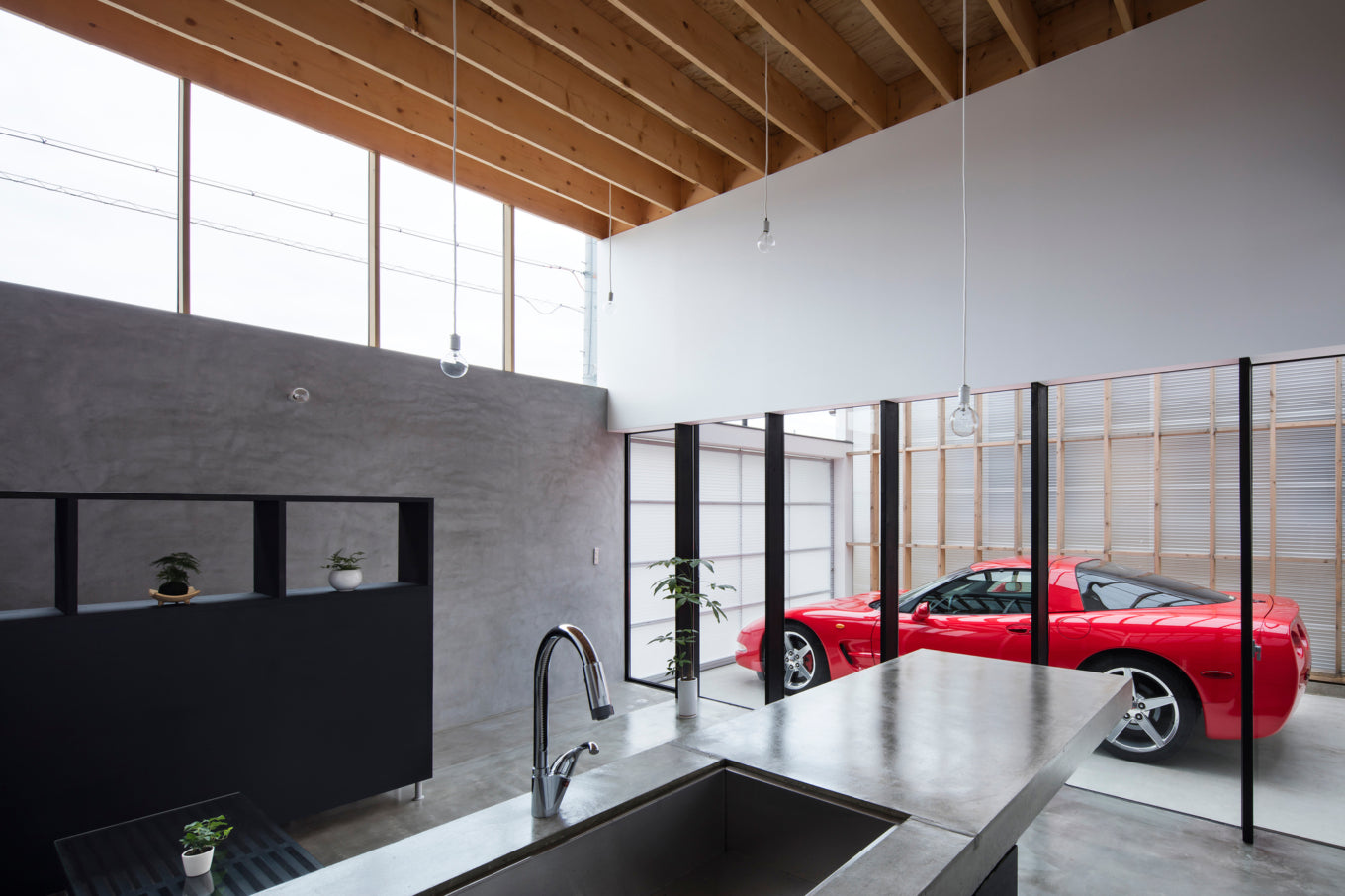 Modern kitchen with concrete island and sink, looking into a garage with a red Corvette C5 sports car.