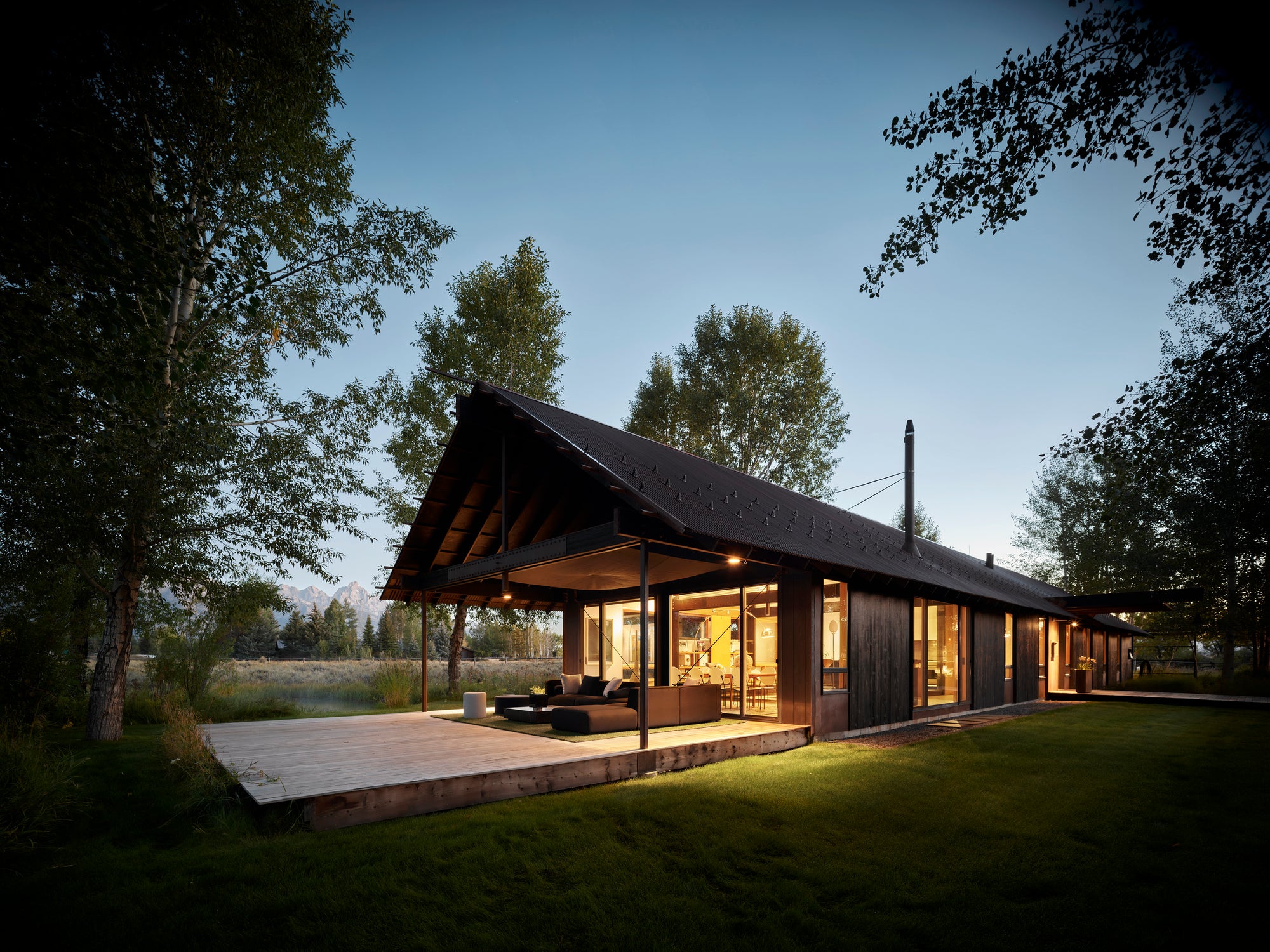 Modern dark wood house, Logan Pavilion, with illuminated interior and deck at twilight, Jackson, WY.