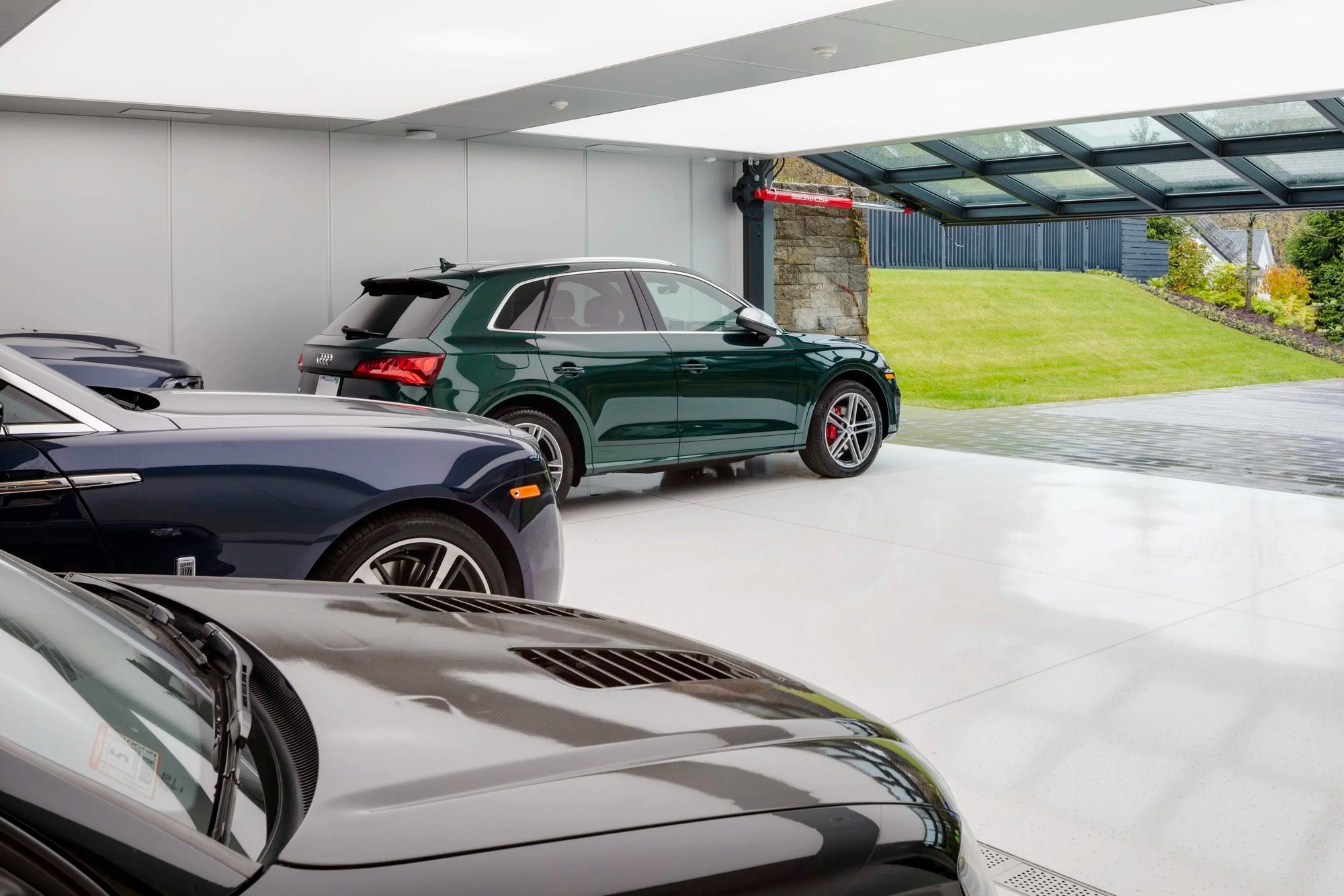 Luxury cars parked inside a modern garage with a view of a garden outside. Cars include a green Porsche SUV and blue Bentley. 