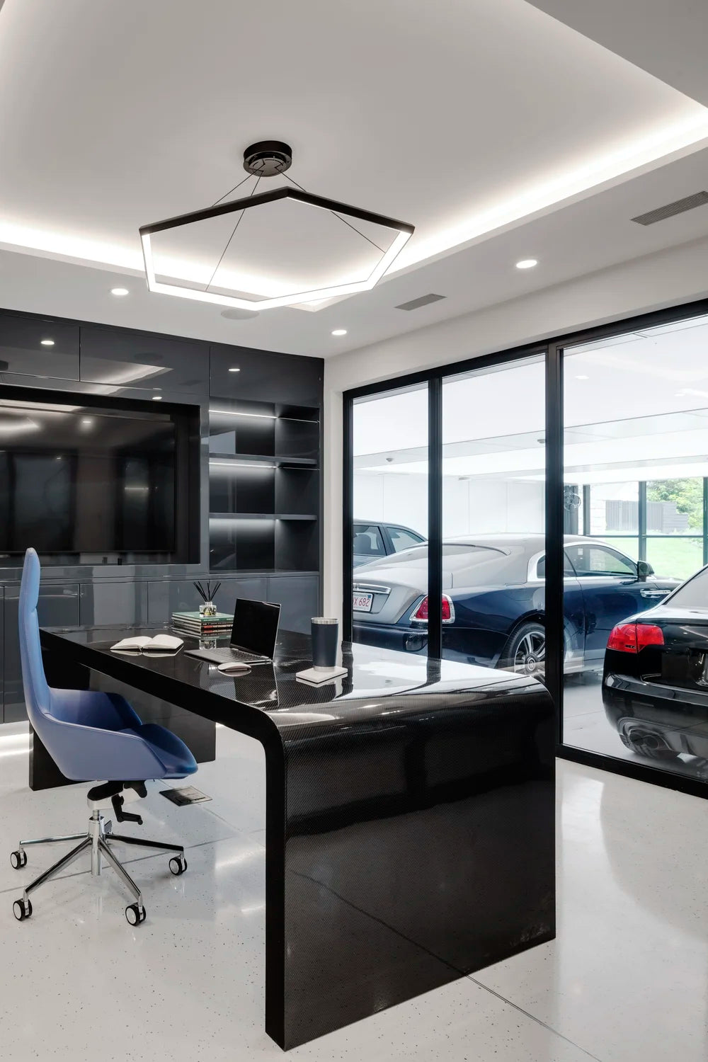 Modern office interior with a black desk, chair, and large windows overlooking car collection in garage. 