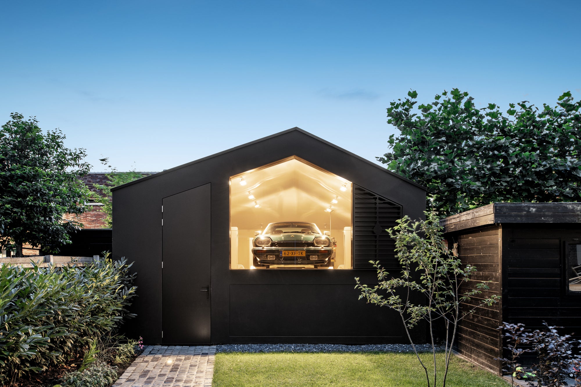 Modern black garage with a classic green Jaguar car inside, surrounded by greenery under a clear blue sky.