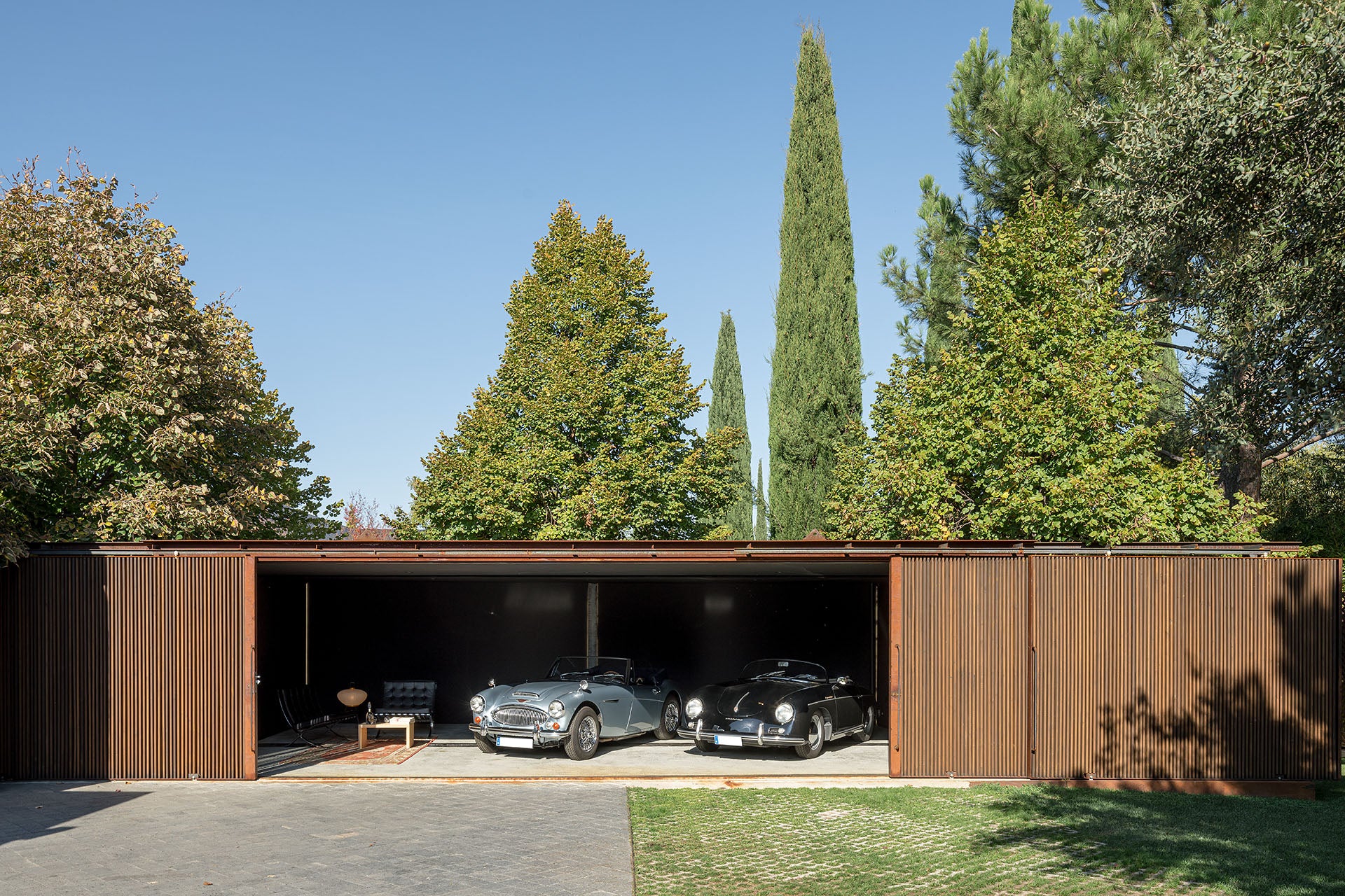 Modern garage architecture with two classic Porsche cars parked inside, surrounded by trees on a clear day.