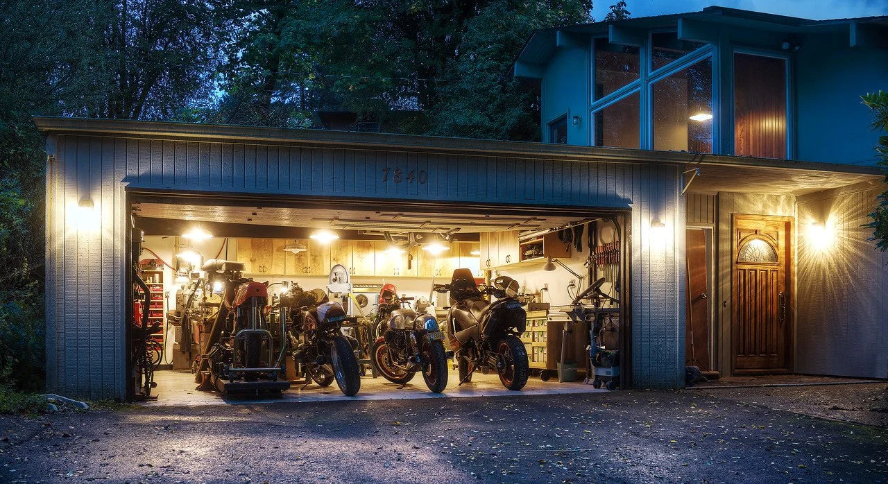 Garage with motorcycles inside, illuminated by exterior lights, surrounded by trees.