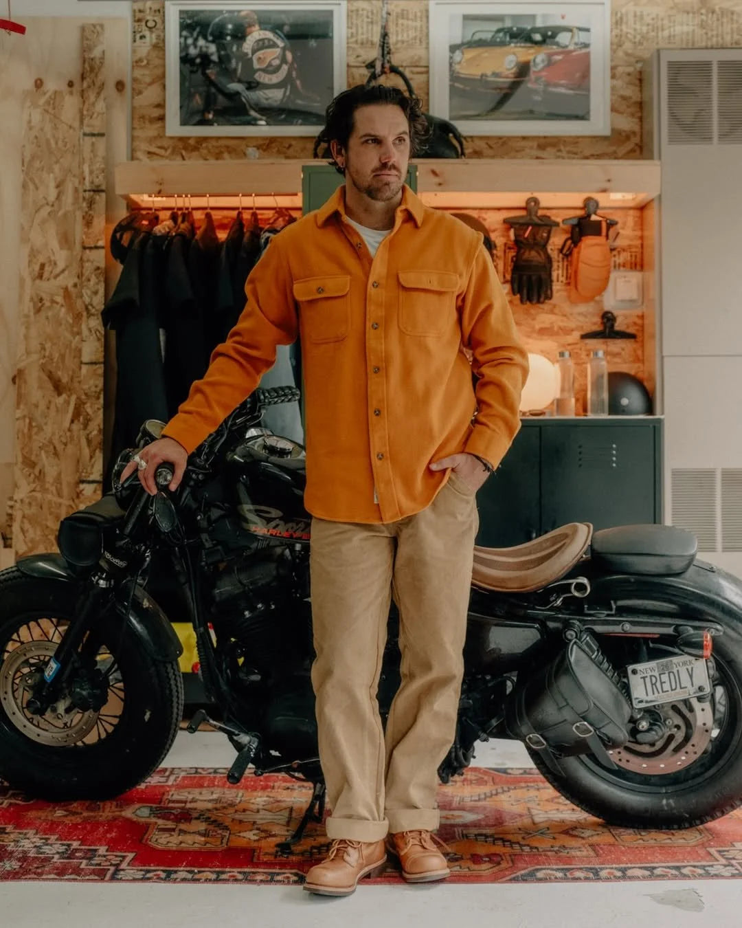 Man in orange flannel shirt and khaki pants stands beside a black motorcycle in a garage.