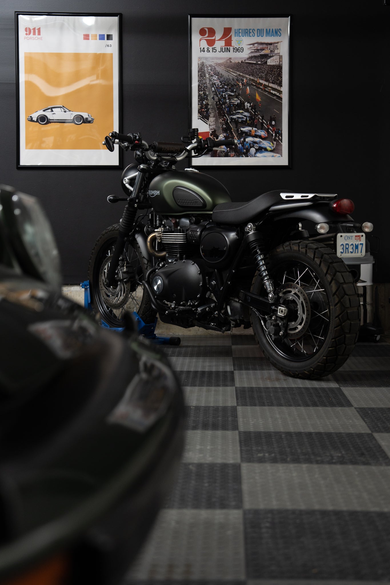 Triumph Scrambler motorcycle parked beneath vintage Porsche and Le Mans posters