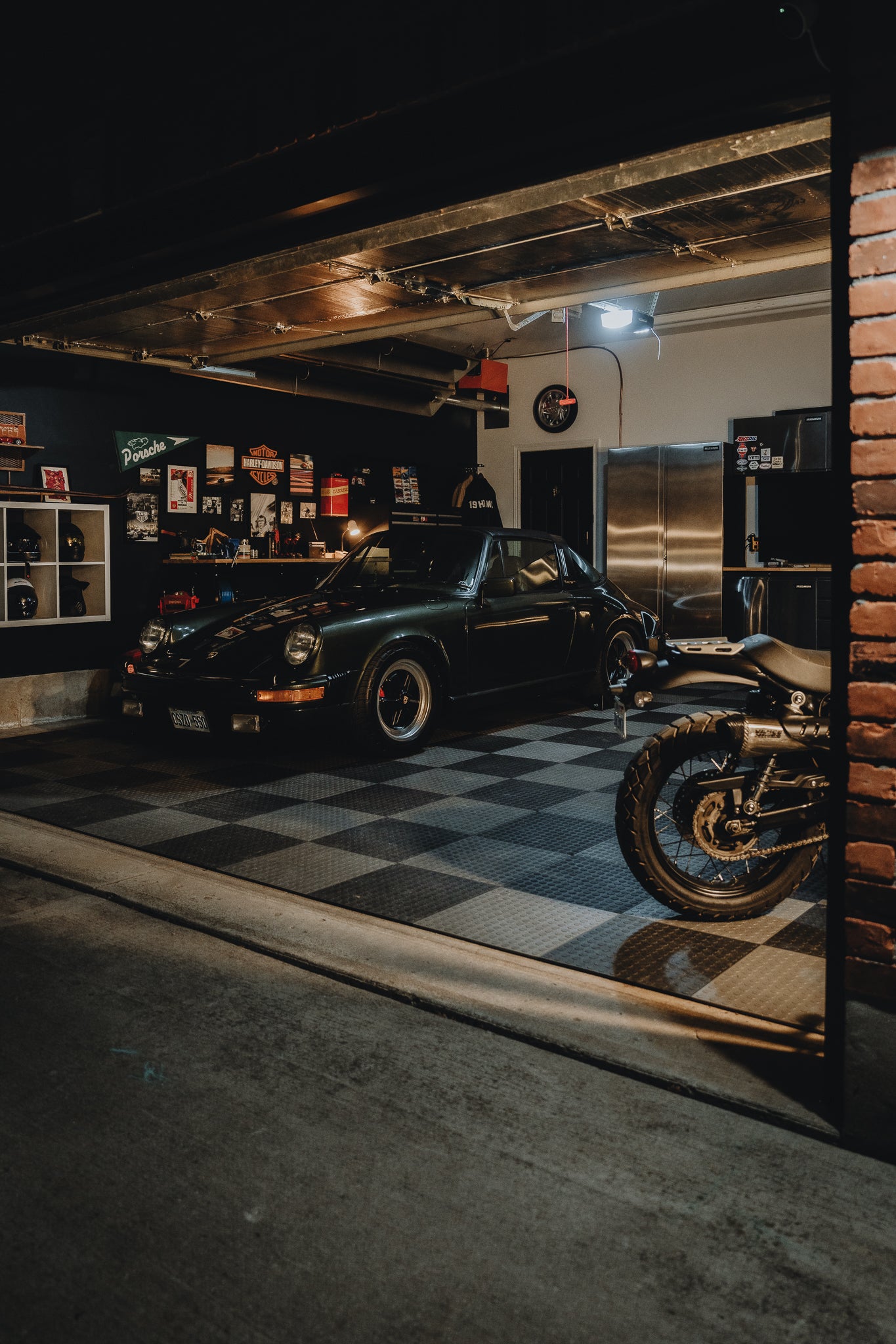 Warm garage interior night shot with a Porsche 911 and motorcycle, checkered floor, and various memorabilia on the walls.