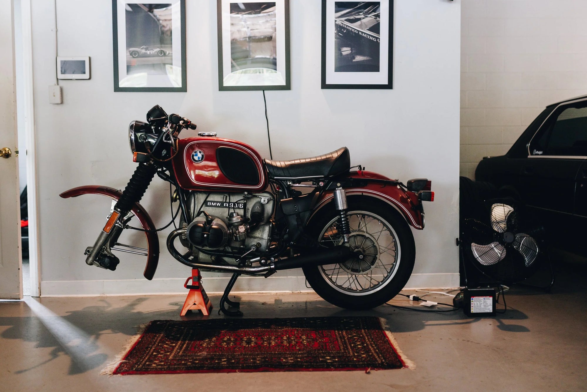 Vintage BMW motorcycle on a stand in a garage with framed pictures on the wall and Persian rug on floor. 