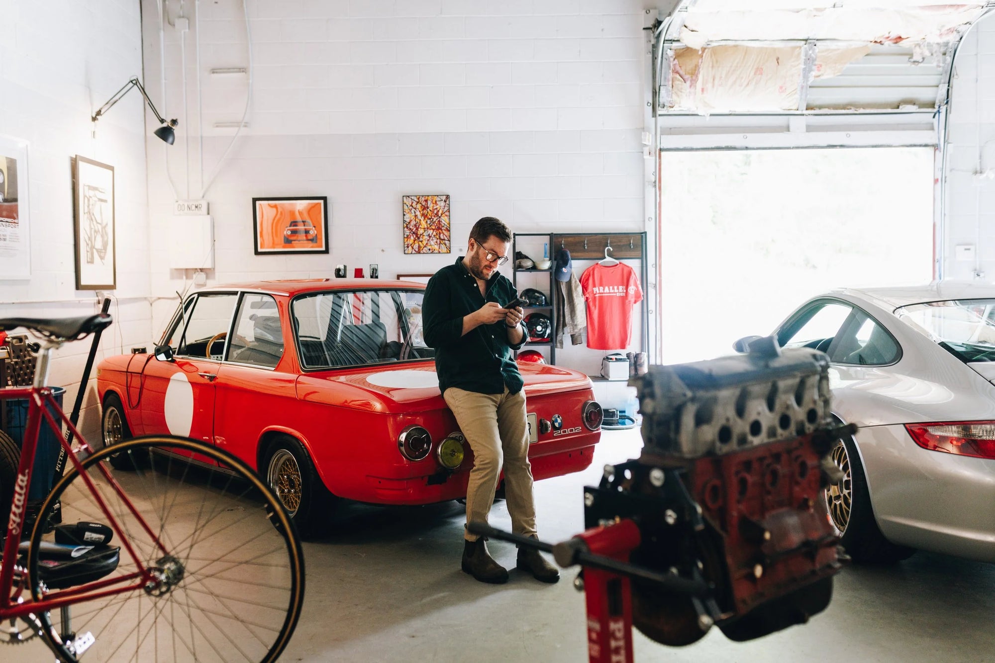 Man checking phone, leaning on a red vintage BMW 2002 in a Charlotte garage with a Porsche and bike.