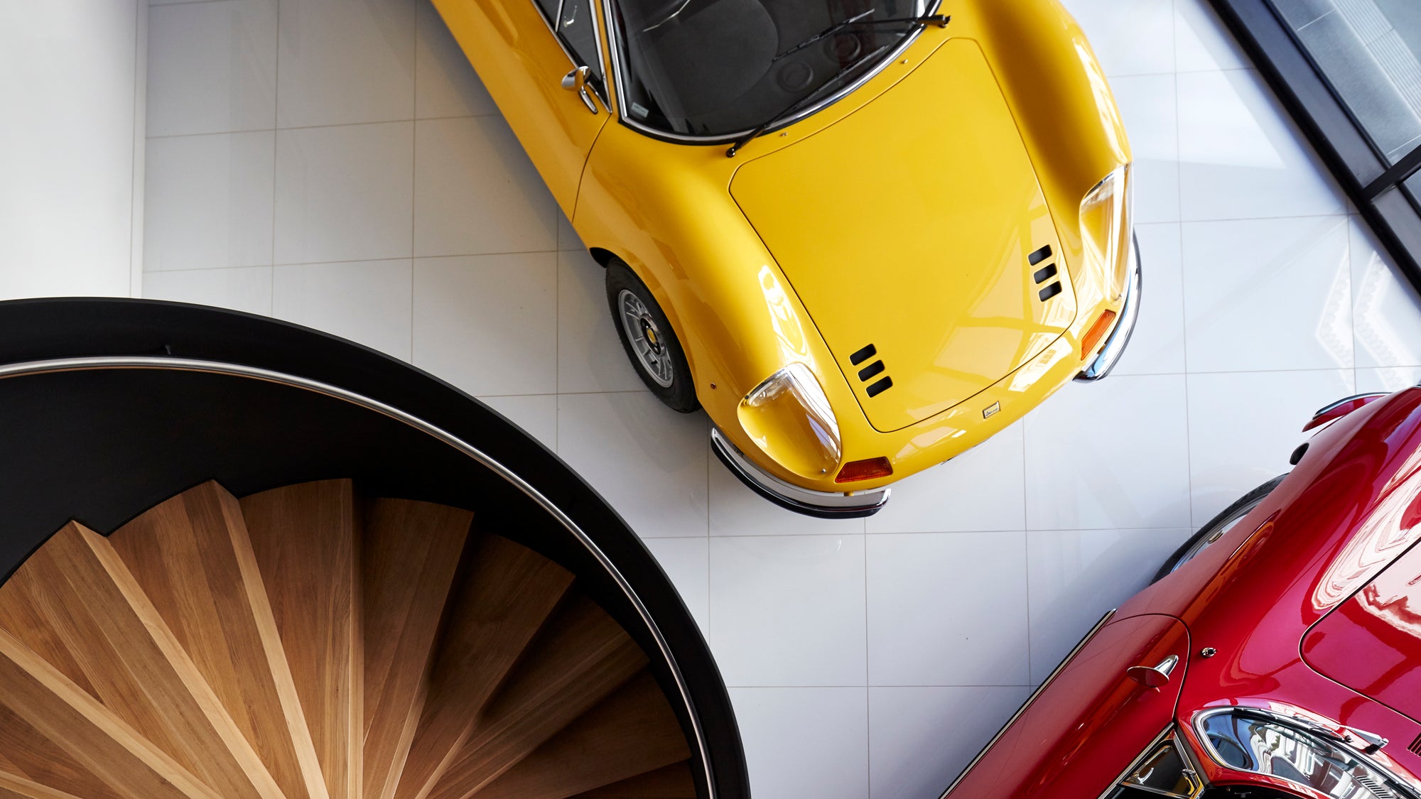Yellow Ferrari Dino and red Porsche on a white floor with a wooden spiral staircase.