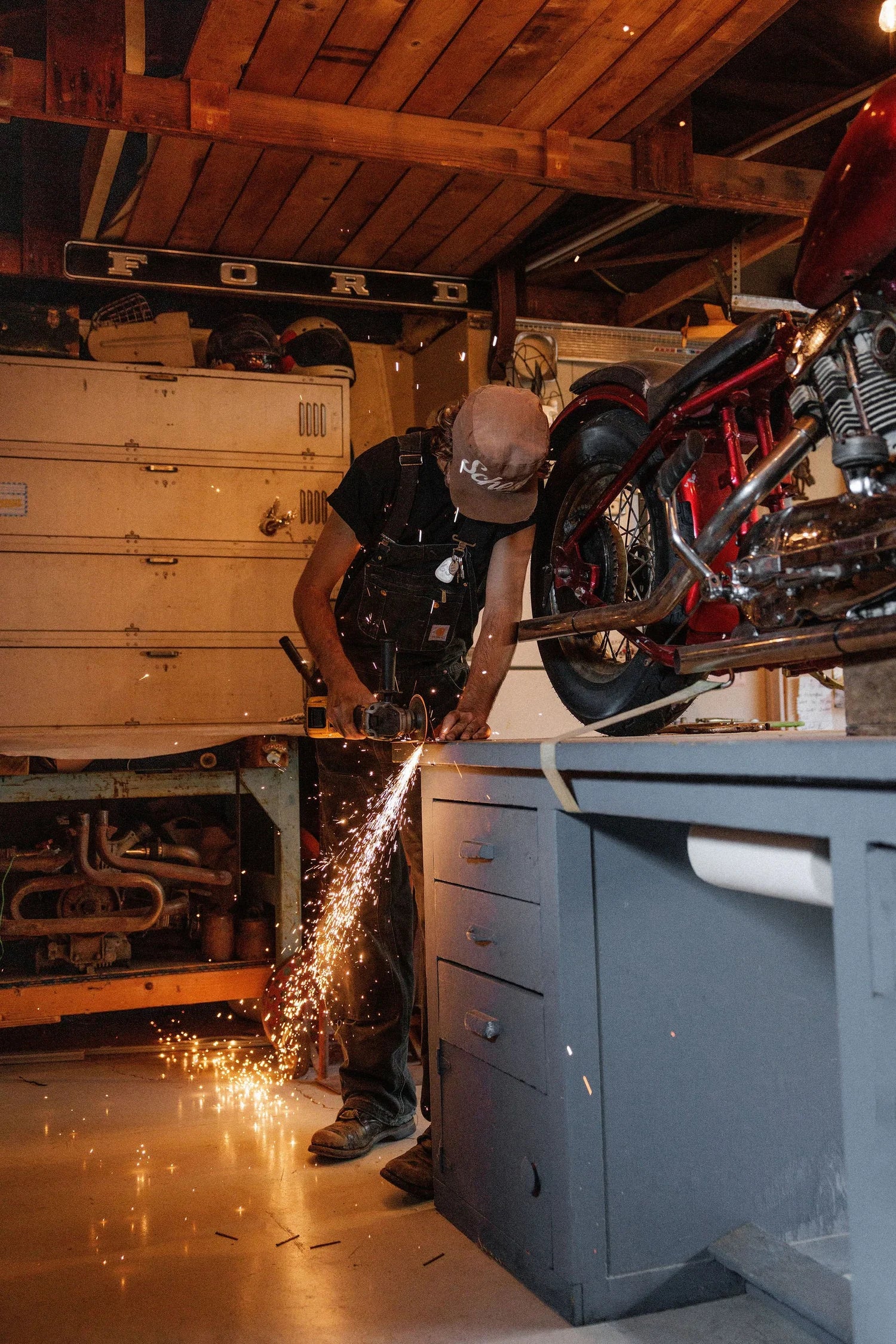 Zander Roe working in his cluttered Northern California garage workshop with tools on his custom motorcycle as sparks fly. 